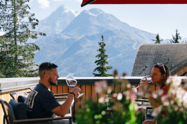 Terrasse du « Terroir des Vignobles » avec un couple qui déguste un verre de vin, face à la montagne, au soleil.